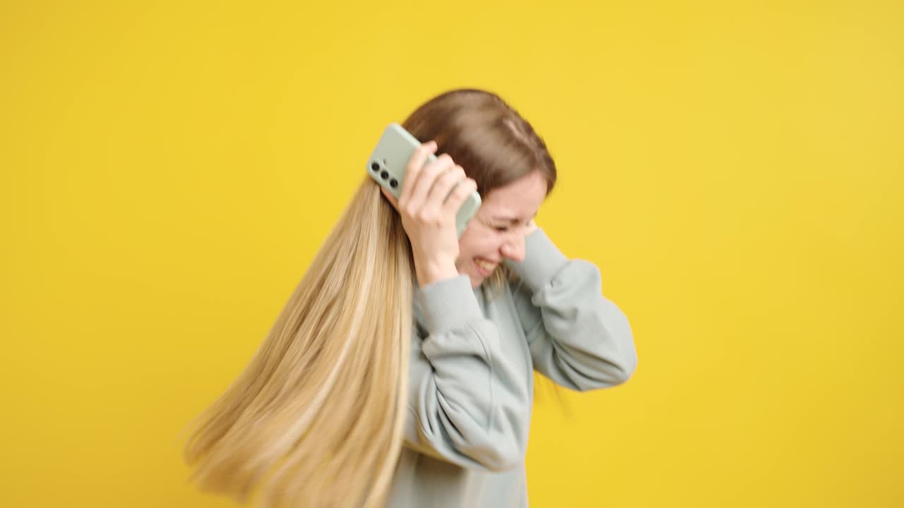 Young woman expressing excitement and laughter with smartphone on yellow background