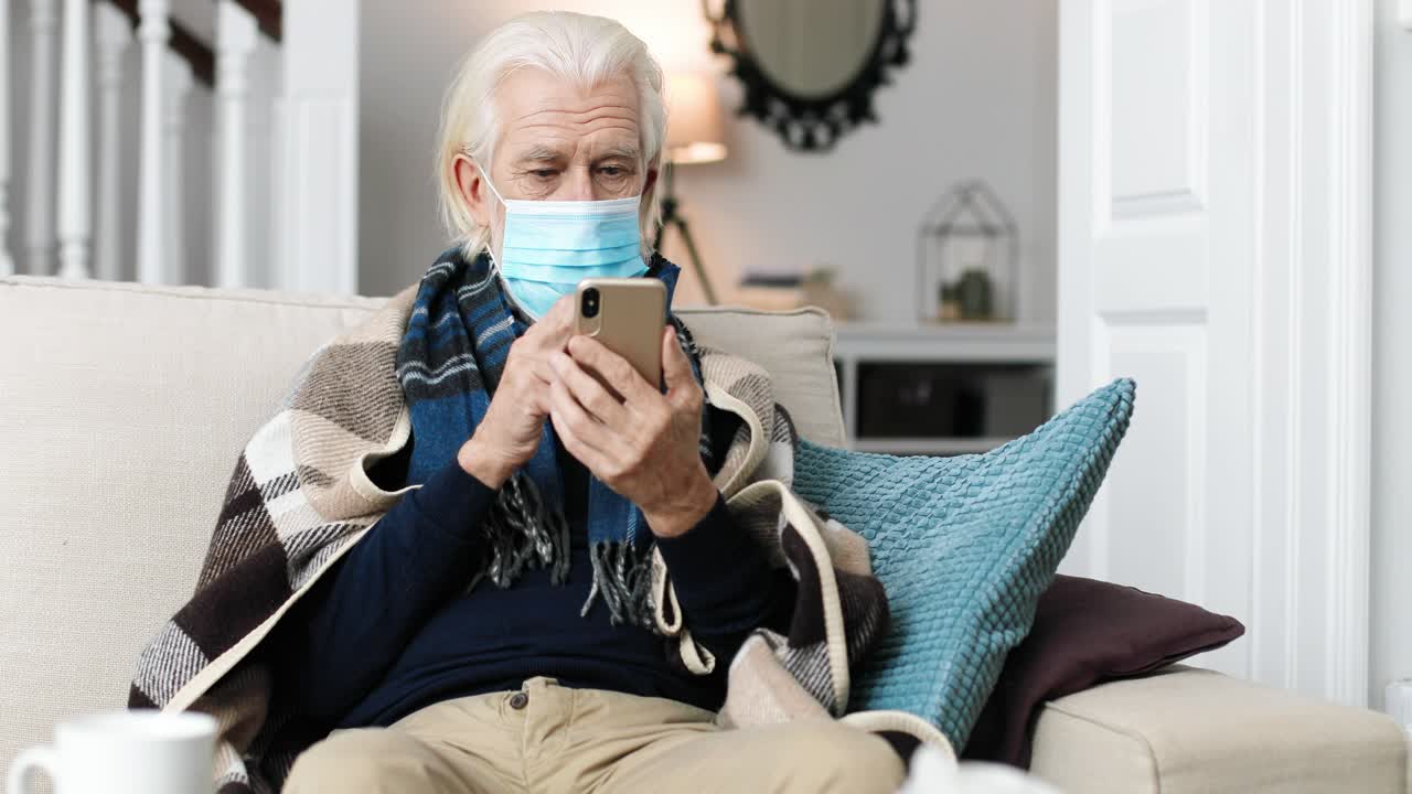 retrato de un hombre enfermo con una máscara quirúrgica sentado en una habitación acogedora y tocando en el teléfono inteligente