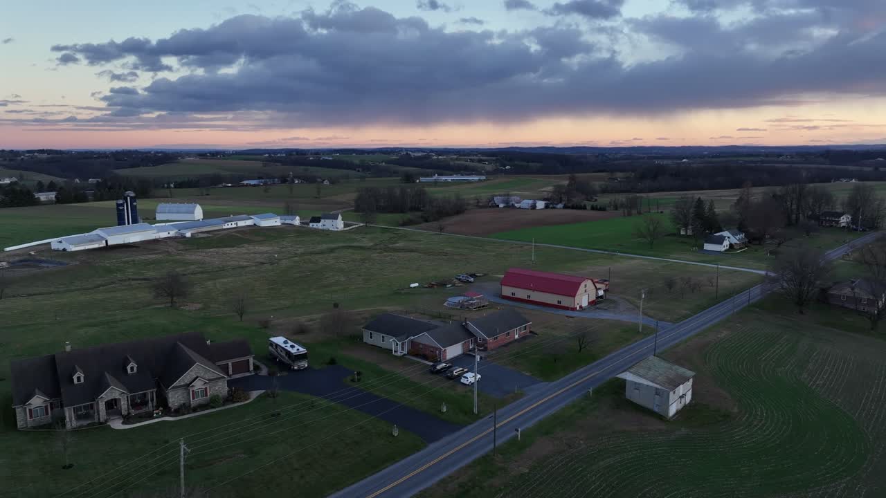 Luftfoto af landligt, amerikansk landbrugsland i skumringen, hvor åbne marker, spredte huse og en rød lade ligger under den falmende himmel. Rolig landlig atmosfære på tværs af det stille amerikanske landskab.