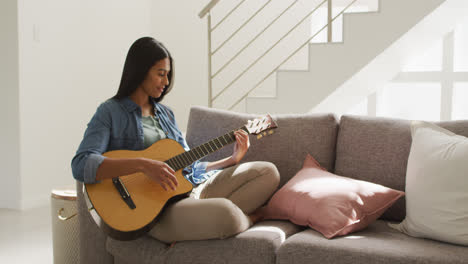 Video-of-happy-biracial-woman-sitting-on-sofa-and-playing-guitar