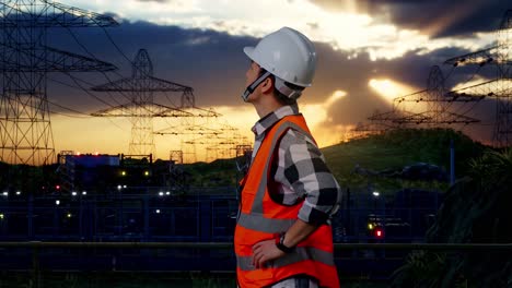 side view of asian male engineer wearing safety helmet looking around while standing with arms akimbo near high voltage tower