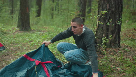 young man smiles while crouched on forest ground, holding tent cover with both hands during camping activity, working alongside partner near tree line with pitched tent visible in background