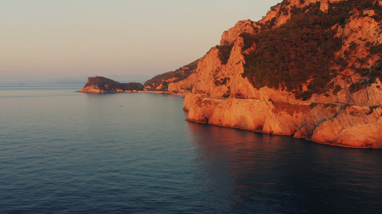 Aerial shot of dramatic, rugged coastal cliffs bathed in warm golden light at sunrise. Winding road along the cliffs, sea reflects the soft gradient of the evening sky. Italy, Liguria.
Capo Noli