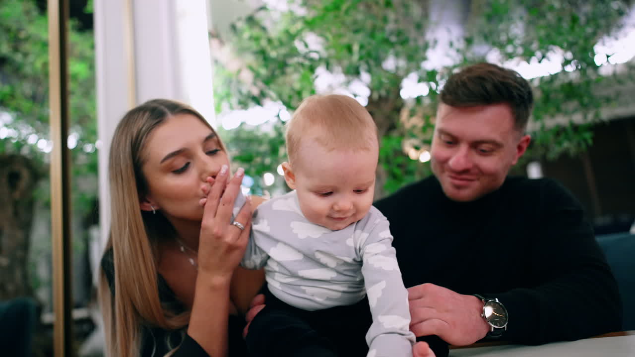 Tiny barefoot infant sits on the table supported by his parents. Mom and dad hold the kid's hands and he tries to stand up.