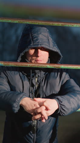 close-up of a person in a hooded jacket resting on an iron bar with clasped hands, illuminated by light, the background features a blurred view of a yellow building and a moving car