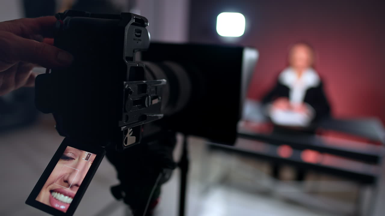Face of a smiling Caucasian beautiful woman on the display of a camera on tripod. Male hands switches a button on the camera. Blurred backdrop.