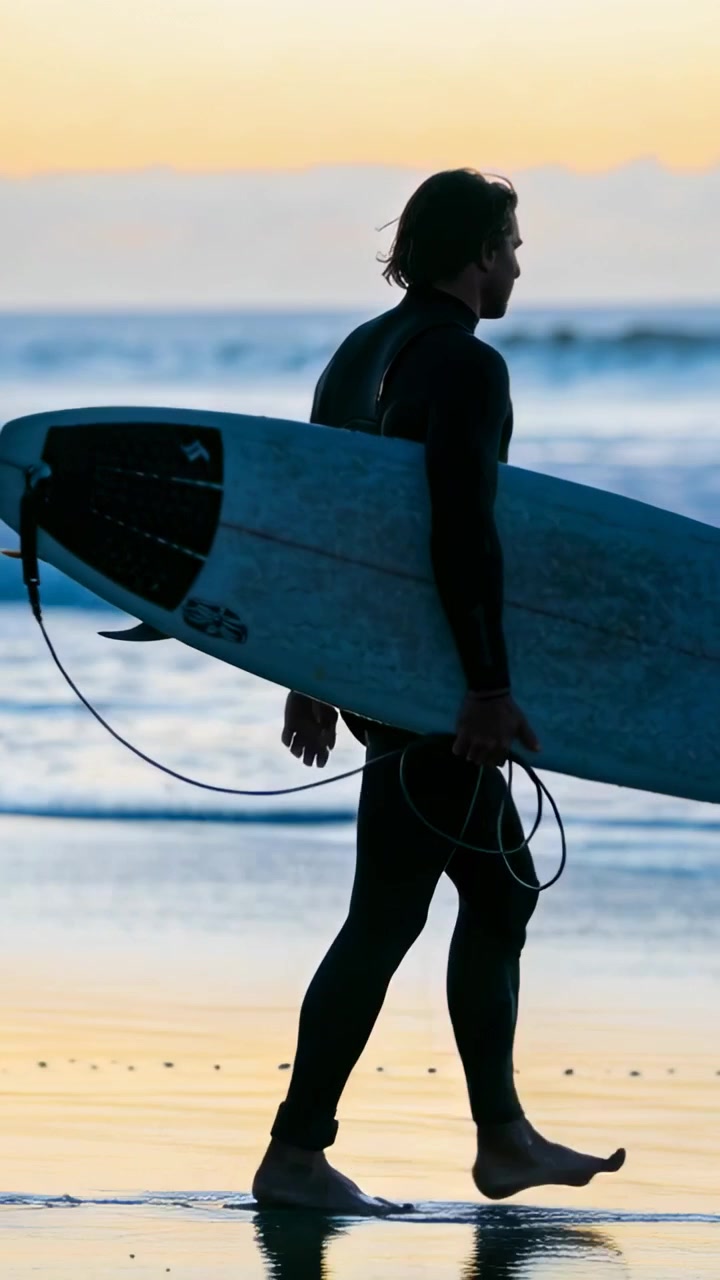 Wideo pionowe: Idący surfer w piance, niosący deskę z leashem po mokrym piasku po surfowaniu, z miejscem na tekst.