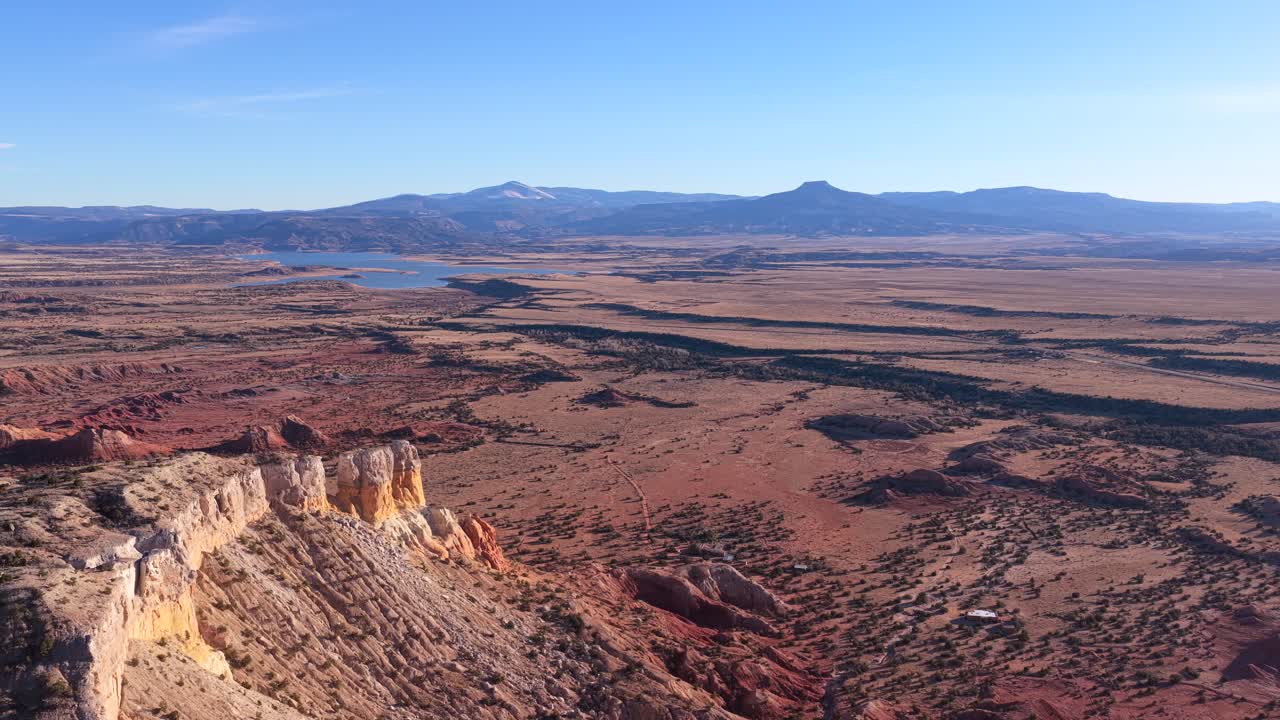 Drone footage glides above red rock cliffs into wide desert plains, revealing layered badlands, sparse vegetation, a distant reservoir, and Cerro Pedernal rising prominently along the horizon