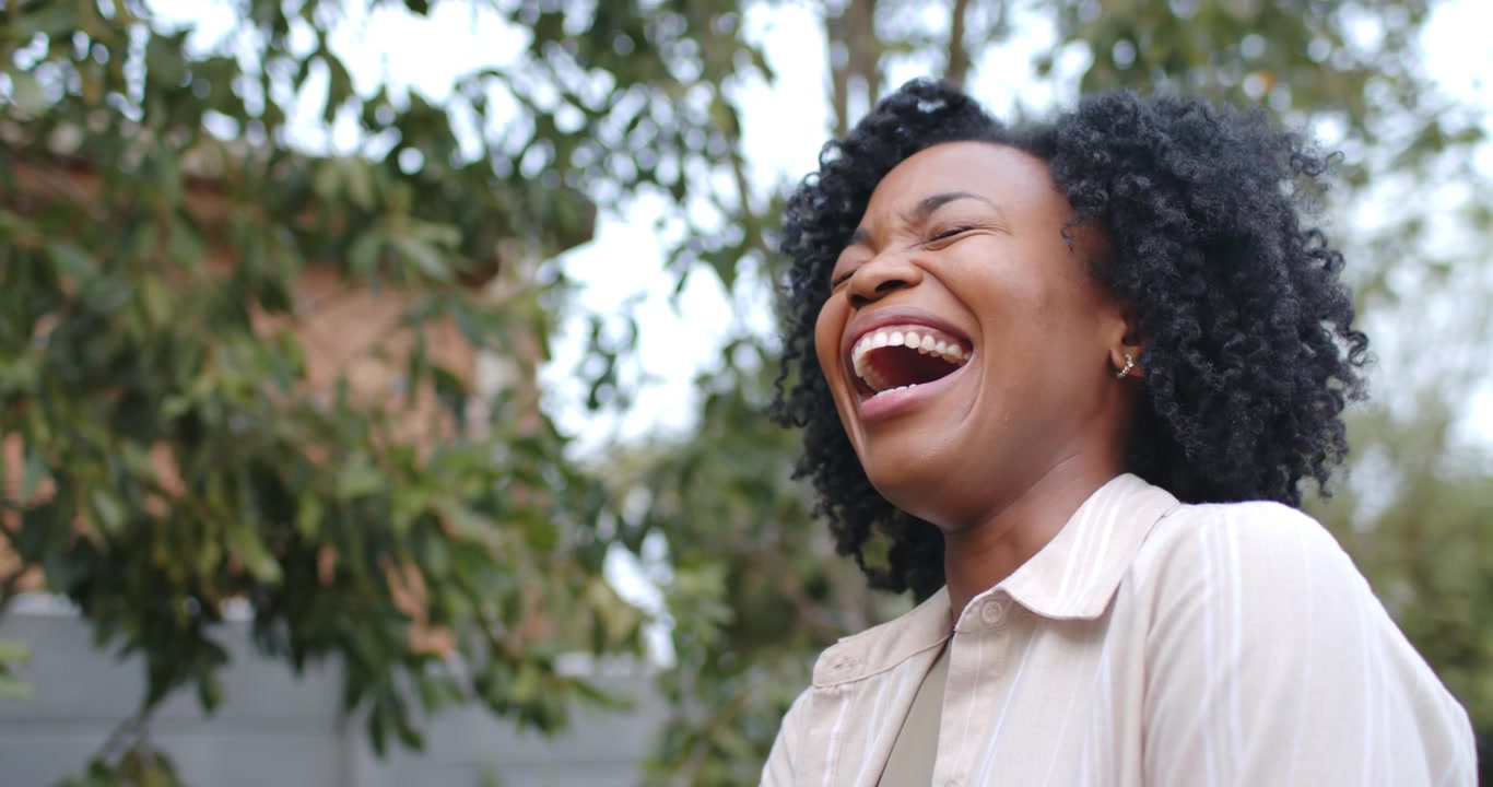 African American woman smiling then laughing, reacting to offscreen voice in yard with bag strap