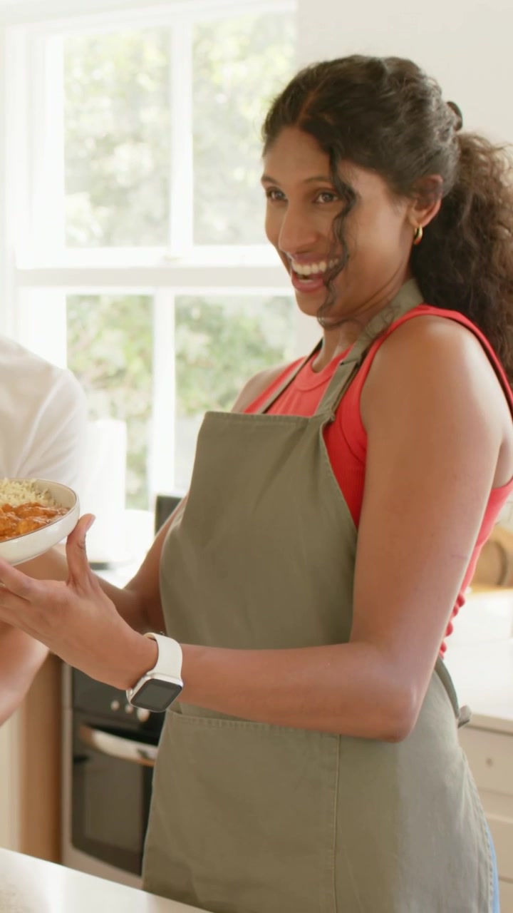 Vertical video: Smiling woman reaching as guest entering, presenting bowl, wearing olive apron