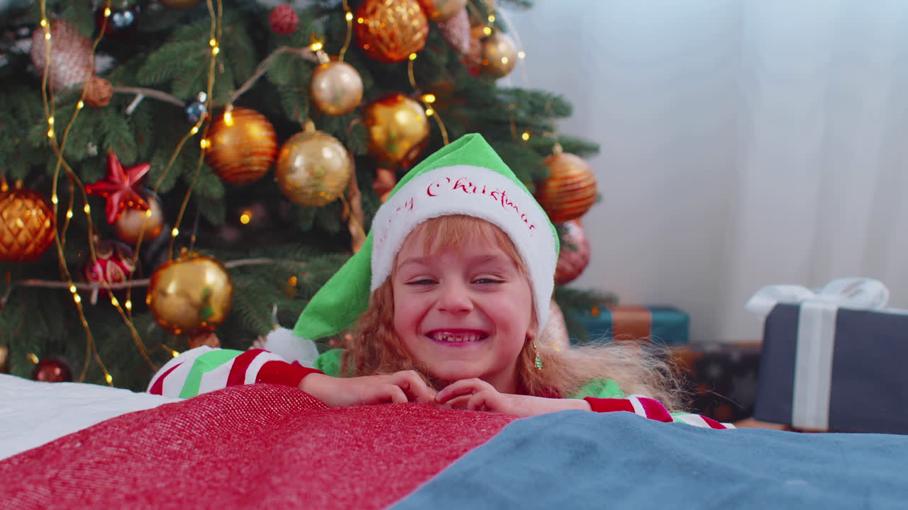 Toddler girl in Santa Claus Elf pajama hiding behind bed, playing hide and seek game, Christmas tree