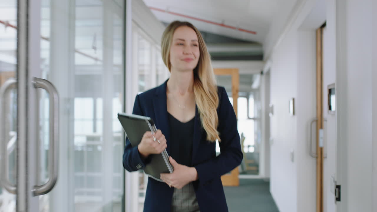 attraente donna d'affari bionda sorridente che cammina in ufficio con un tablet computer che gode di una carriera di leadership di successo nel posto di lavoro aziendale 4k