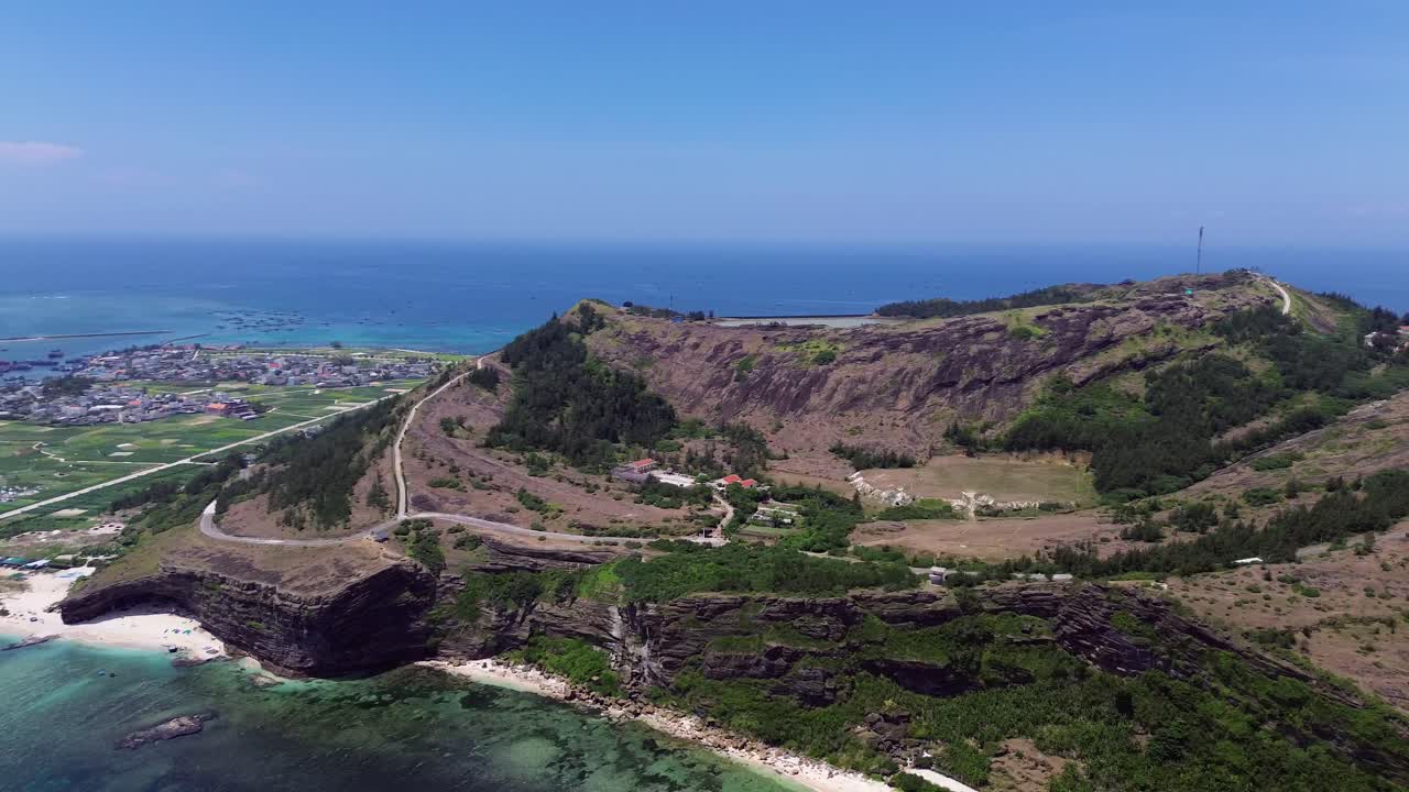 Drone pan right showing layered rock formations, tropical vegetation and deep blue ocean on a sunny day