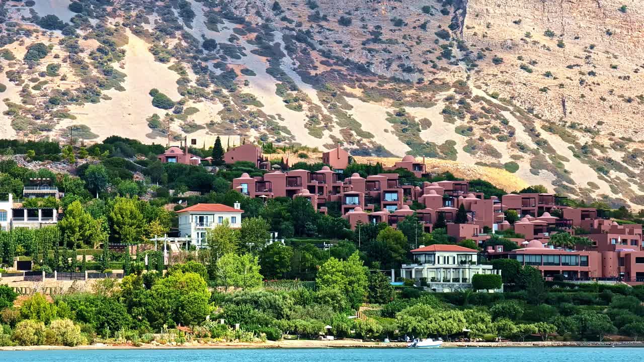 Sudan Elounda limanının manzarası, sahil binaları, yamaç villaları ve kayalık bir arka plan ile. Akdeniz tatil atmosferi sunan manzaralı bir kıyı destinasyonu görüntüsü.