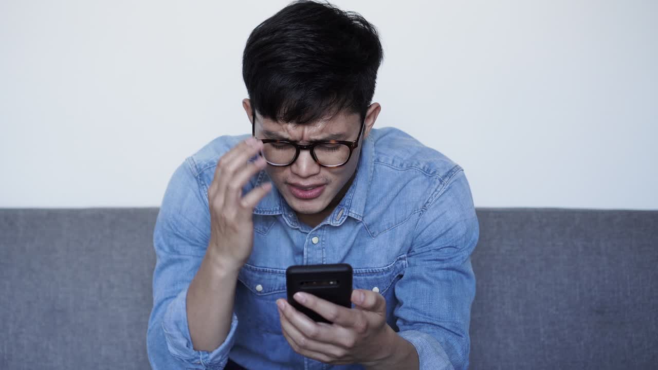 가까운 장면: 젊은 아시아 남자가 진지한 감정으로 얼굴을 어보고 소셜 미디어, 웹 사이트 또는 블로그에서 스마트 폰에서 논평을 읽거나 가짜 뉴스를 읽습니다.