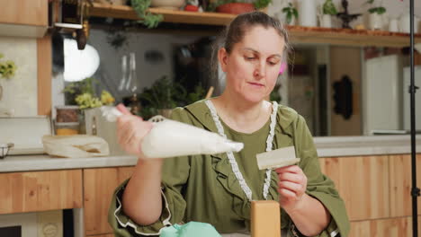 housewife in green dress and apron prepares for baking by dropping icing on baking base, placing paper on it, then positioning pink cookie carefully while sitting in cozy home kitchen