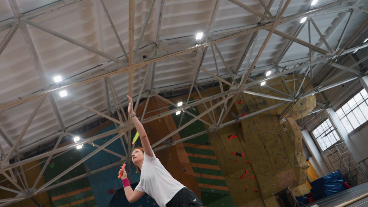 Schiacciata di badminton dall'alto di un giocatore bianco su un campo al coperto, follow-through esplosivo e salto elevato, espressione concentrata e polsino in evidenza, luci del tetto della palestra e sfondo di parete da arrampicata, dinamico.