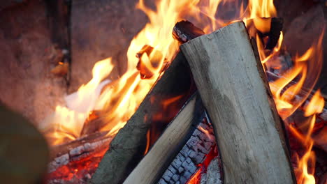 static fhd shot of logs and papers burning in a campsite fire with flames all around, charred embers glowing below and rocks in the background