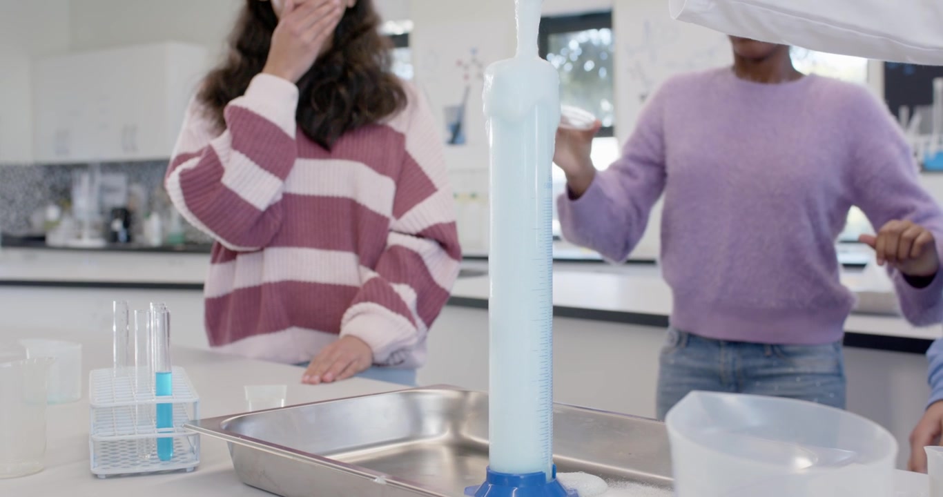 Studentesse adolescenti di diverse etnie che indossano occhiali protettivi, versano un reagente tramite un imbuto in un cilindro graduato in laboratorio per una dimostrazione.