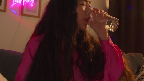 집 파티에서 알코올을 마신 후 소파에 앉아있는 여성은 숙취를 위해 물 한 잔을 마시고 있습니다.