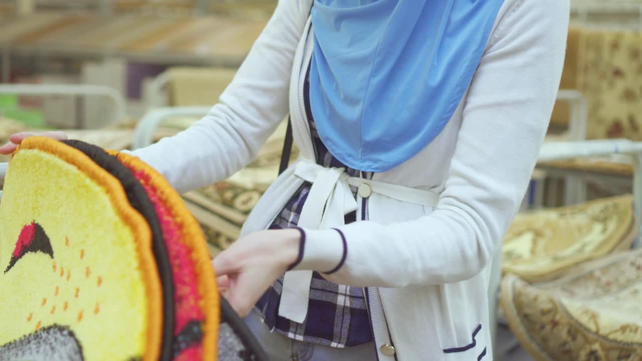 Young muslim woman in a hijab and glasses in a carpet store chooses a carpet