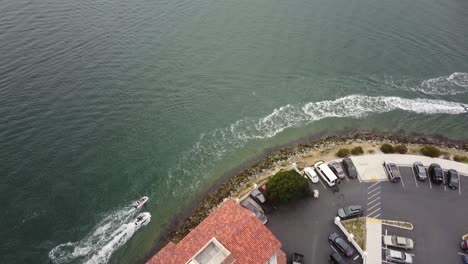 Drone-shot-looks-down-on-two-boats-speeding-past-a-building-and-parking-lot-on-the-harbor-and-pans-up-to-reveal-the-North-San-Diego-Bay,-California