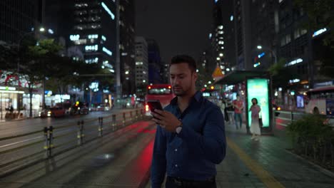 Joven-Atractivo-Hombre-Barbudo-Parado-En-La-Parada-De-Autobús-Esperando-El-Transporte-Por-La-Noche-Después-Del-Trabajo-Y-Usando-El-Teléfono-Para-Encontrar-La-Hora-De-Llegada