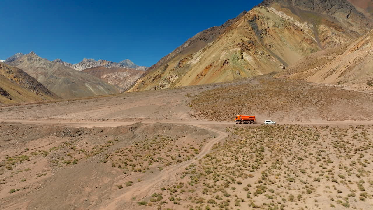Orange mining truck driving on dirt road in arid valley, colorful Andes mountains under clear blue sky, Chile. Aerial sideways