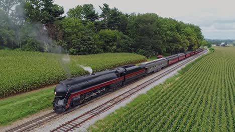 a drone view of an antique restored steam engine and passenger coaches getting ready to leave a small train station on a summer day