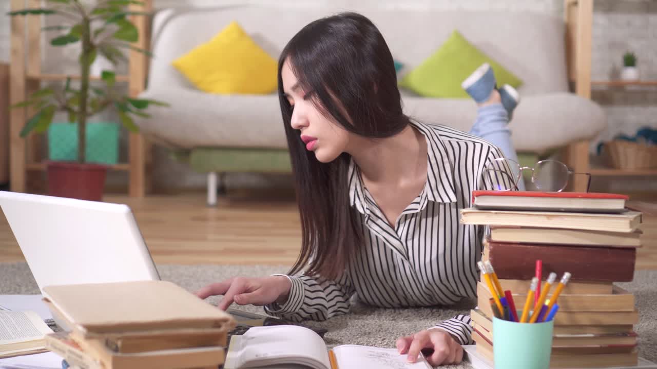 아시아 학생 은 노트북 과 책 을 가지고 거실 의 바닥 에 누워 시험 을 준비 하고 있다