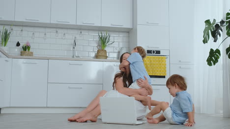 Loving-mother-and-children-hugging-and-having-fun-playing-sitting-on-the-floor.-Fun-and-happy-family.-The-game-after-viewing-the-laptop