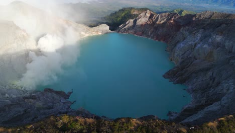 right-orbit drone shot circles kawah ijen, revealing its vivid blue-green acidic lake, smoking sulfur vents and rugged caldera rim high in the mountains of east java, indonesia