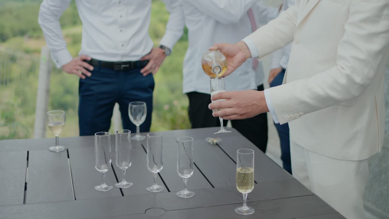 Groom in a white suit pouring champagne into glasses on a table, with friends gathered around for a celebration