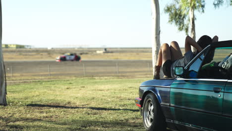 pareja sentada en el capó del auto y viendo la carrera de autos durante el día soleado