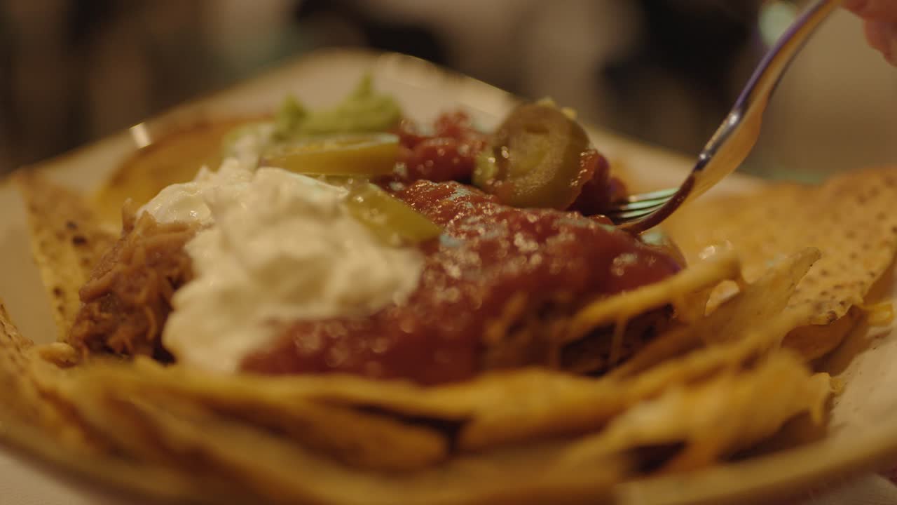 Close-up of nachos with salsa, cream, jalapeños; hand scooping under warm, ambient bar lighting
