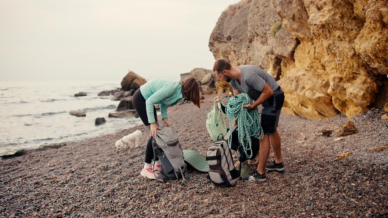 A group of rock climbers, two girls and a brunette guy came to the rocky seashore and lay out their backpacks in order to put on climbing equipment near the yellow rocks on the sea during the day