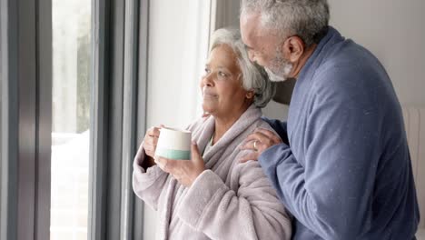 Feliz-Pareja-Birracial-Senior-Mirando-Por-La-Ventana-Y-Abrazándose,-Inalterada,-En-Cámara-Lenta