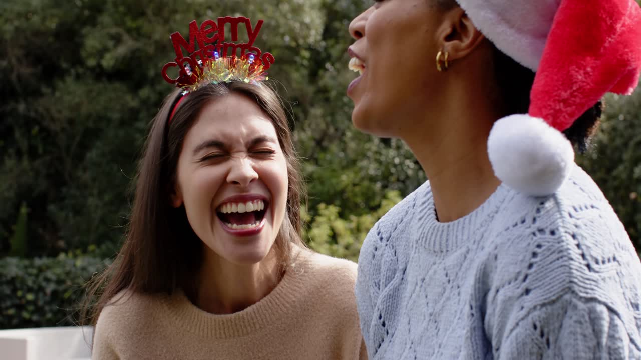 Diverse female friends reacting to headband, Santa hat by planting kisses and laughing on bench