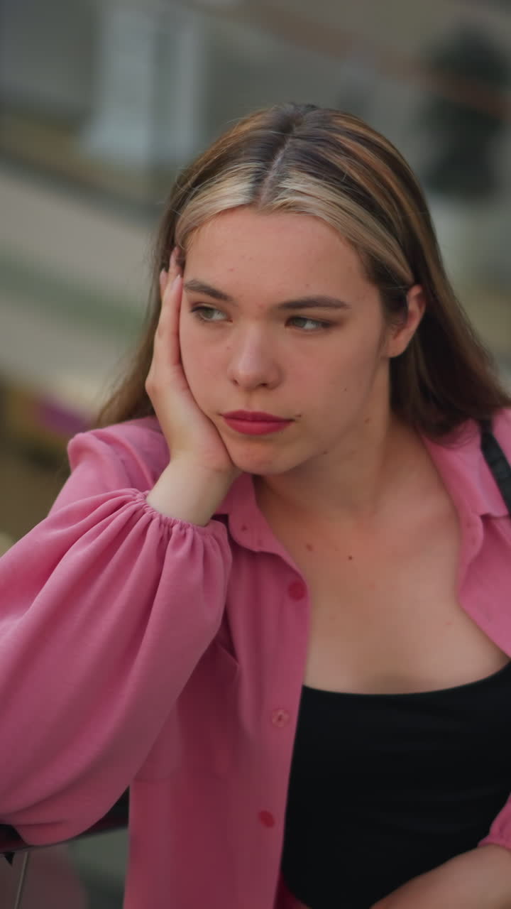 분홍색 옷을 입은 여자가 그녀의 에 손을 놓고, 사려 깊게 보이며, 오른손에 쇼핑 가방을 들고, 쇼핑몰의 흐릿한 배경으로 움직이는 에스컬레이터를 타고 있습니다.