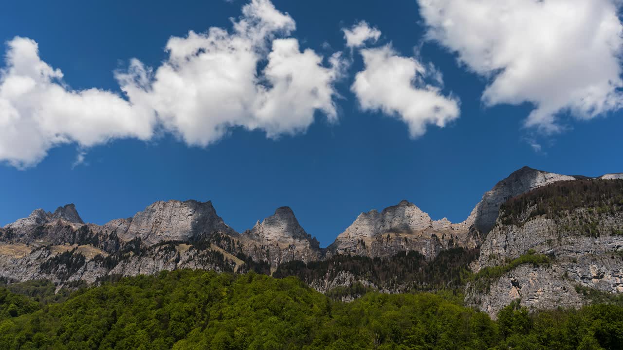 Time-lapse. Clouds swirl over the Churfirsten mountain range. The shadows of the clouds dance on the mountain peaks. The rocks are illuminated by the sun. It's a spring day with a natural landscape.