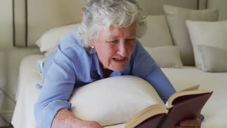 mujer mayor caucásica feliz en casa disfrutando de leer un libro, acostada en la cama en un dormitorio soleado