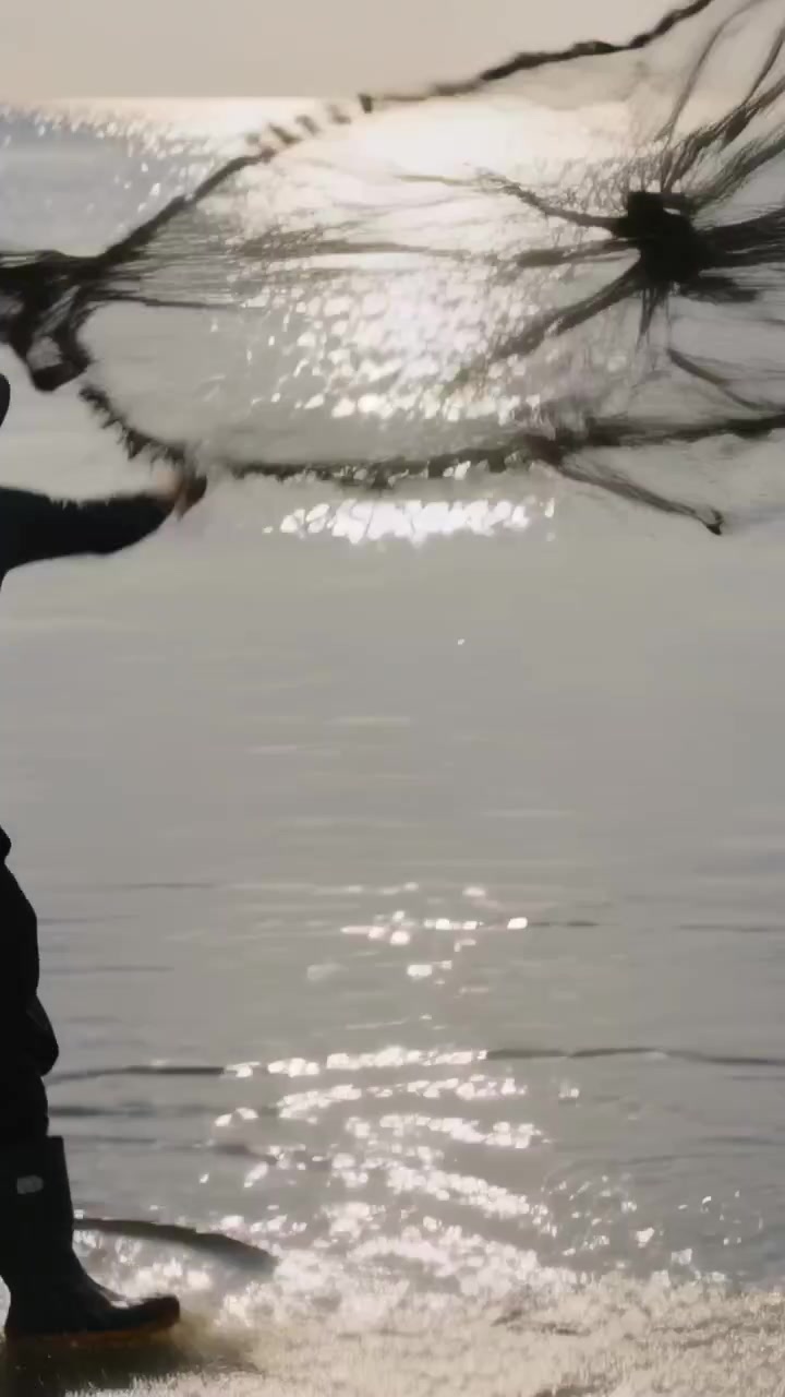 Video verticale: Uomo che cammina tra le onde, prepara e lancia una rete da pesca, con cappello e stivali, spazio per il testo