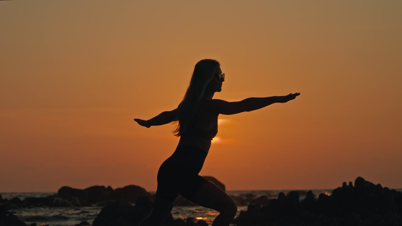Gün batımında gökyüzünde güzel bir turuncu parıltı varken, arka planda sakin dalgalar görünürken, savaşçı yoga pozisyonunda eğilen siluet bir kadın.