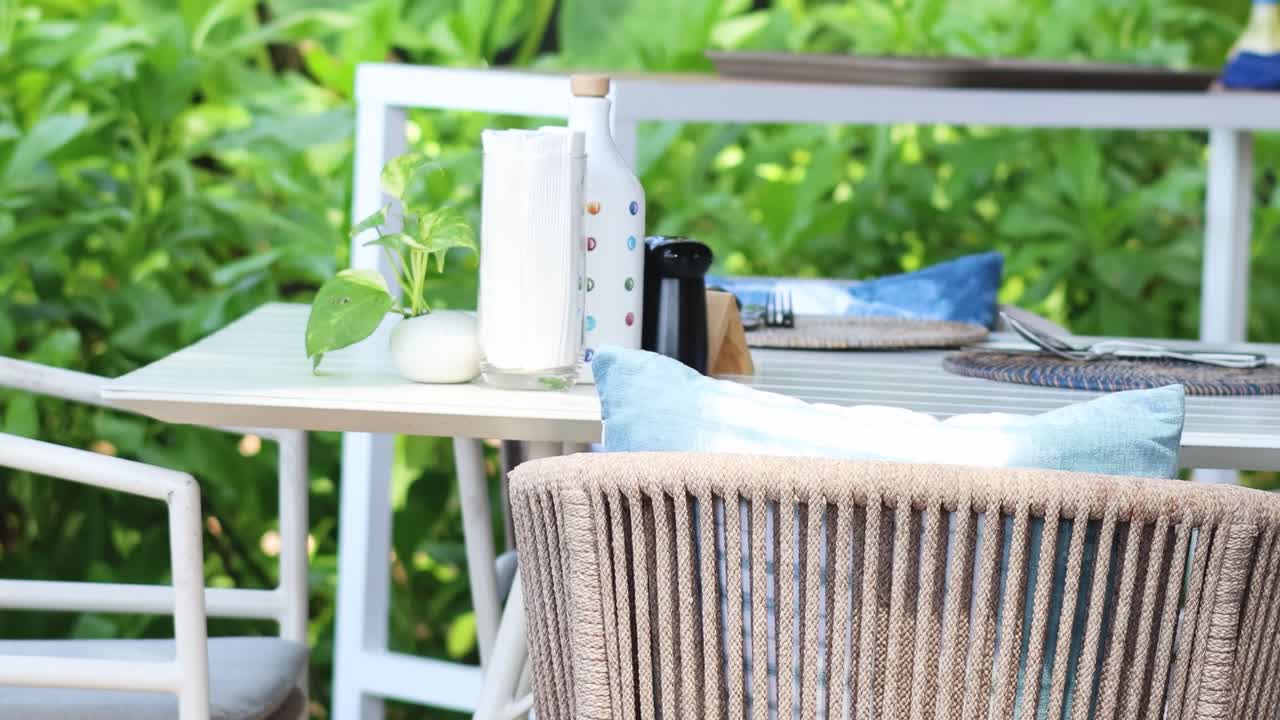 A comfortable patio chair and table set against lush green foliage, featuring decorative cushions and a small plant.