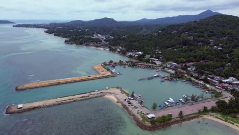 aerial drone footage showing baan tai pier and the surrounding coastal town on koh phangan island, surat thani, thailand, with tropical hills, beach shoreline, and blue sky