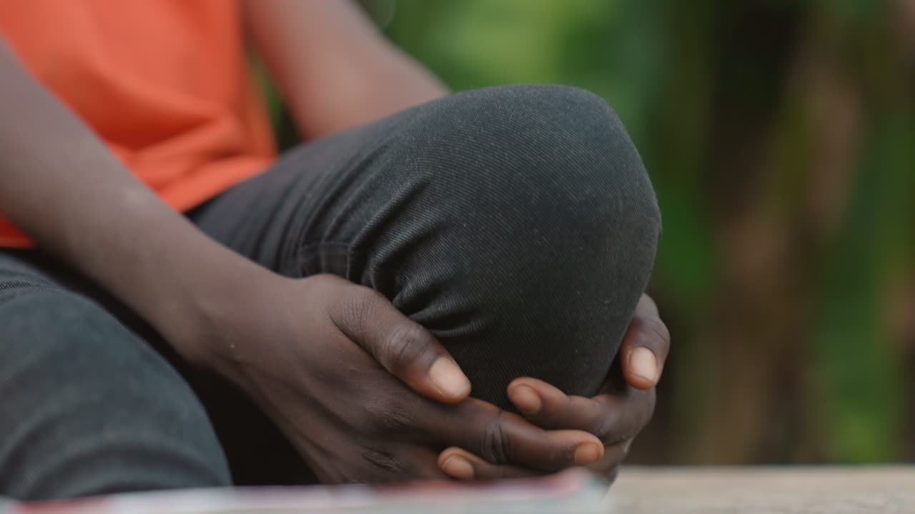 Black athlete clutching knee after twist on outdoor bench wearing black sport leggings and orange top, hands gripping knee in pain, tense closeup showing fabric texture and melanin skin detail, moment