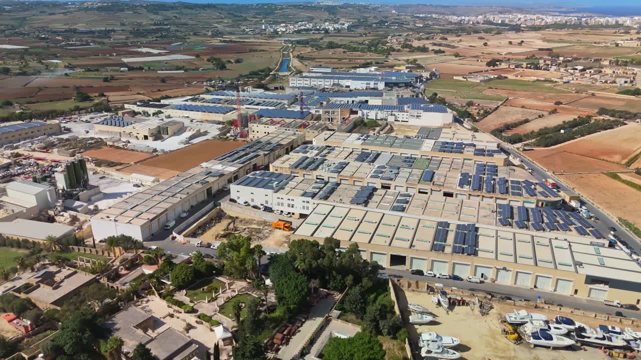 Aerial view showing the large industrial and warehouse complex near Naxxar, Malta, with solar panels on rooftops, surrounding agricultural fields, and distant coastal horizon under a clear blue sky