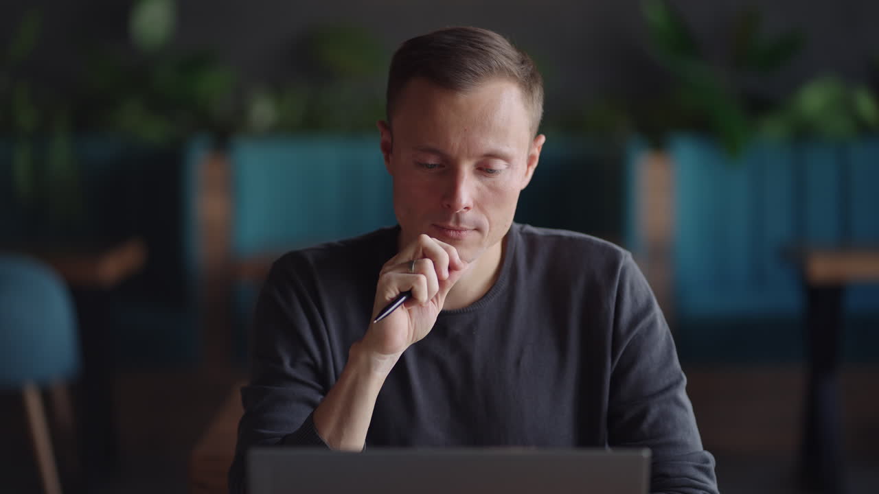 Close-up portrait of ponder young man in office. Designer plans his work and holding a pencil in his hand. Shooting is slow motion from below. Thoughtful serious man sit with laptop thinking solution