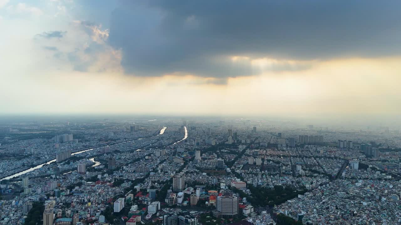 Aerial view over Ho Chi Minh City (Saigon), Vietnam, during sunset, as dramatic clouds cast shadows across the dense urban sprawl. A moody and cinematic atmosphere of a Southeast Asian metropolis.