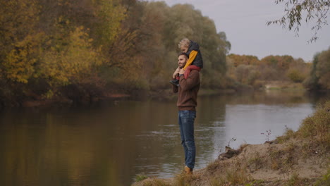joyful man is holding child boy on shoulders and enjoying nature of river and forest at autumn happy father and son at weekend walk picturesque fall landscape
